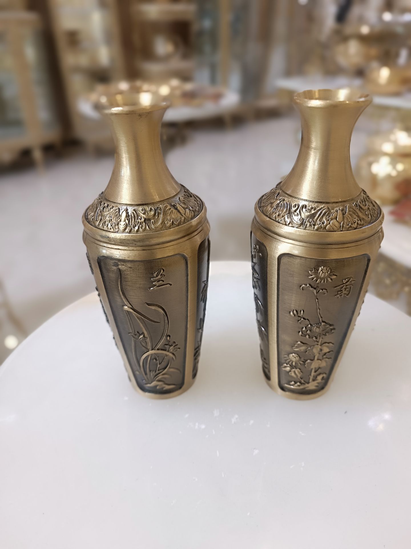 دو عدد گلدان برنجی با رنگ آنتیک و حکاکی‌های ظریف طرح گل و گیاه روی میز سفید فروشگاه متالوکس